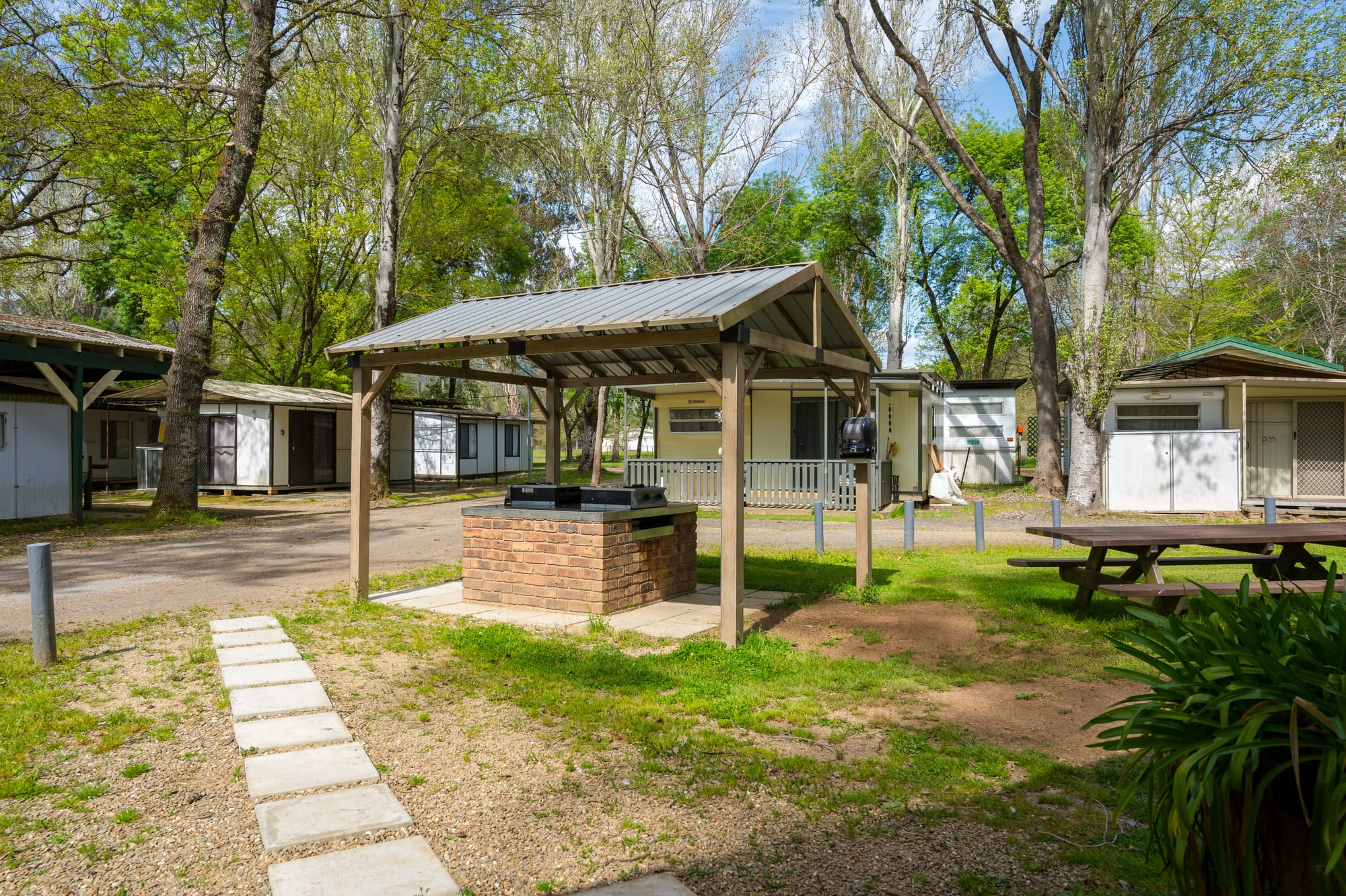 Covered brick BBQ area with paved pathway and mature trees at Bright Accommodation Park, Bright VIC