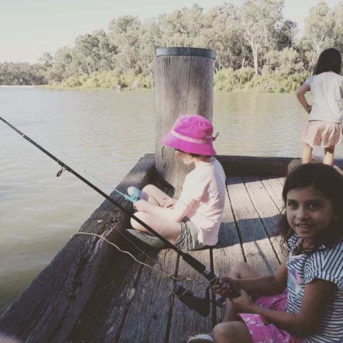 Children fishing off a timber jetty on the river at Torrumbarry Weir Holiday Park, Patho VIC, with fishing rods and gum trees behind