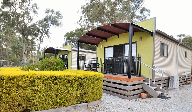 Green and cream cabins with curved awning and deck at Torrumbarry Weir Holiday Park caravan park, Patho VIC, set among gum trees
