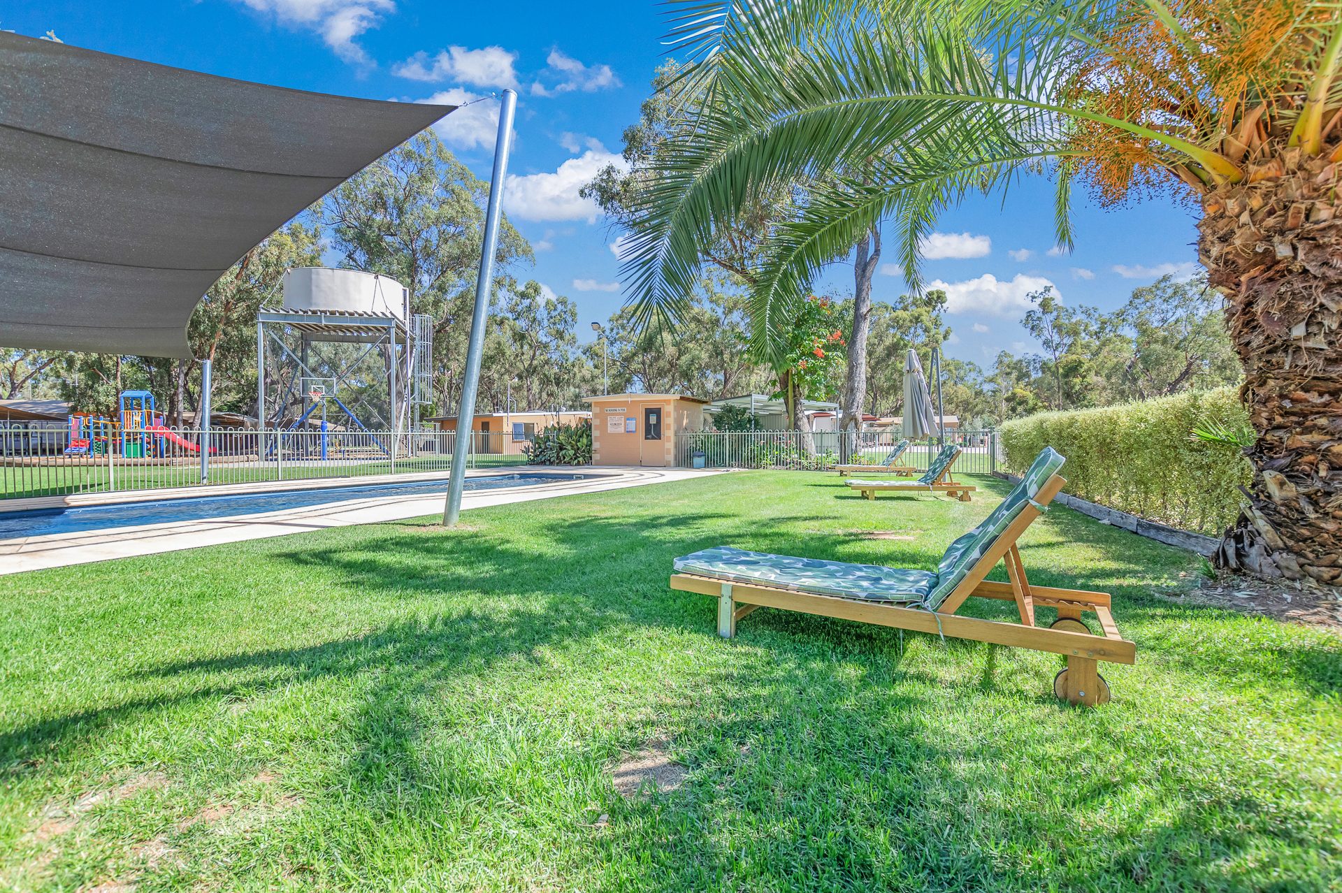 Timber sun lounger with green cushion on the lawn beside the pool at Torrumbarry Weir Holiday Park, shade sail and playground nearby