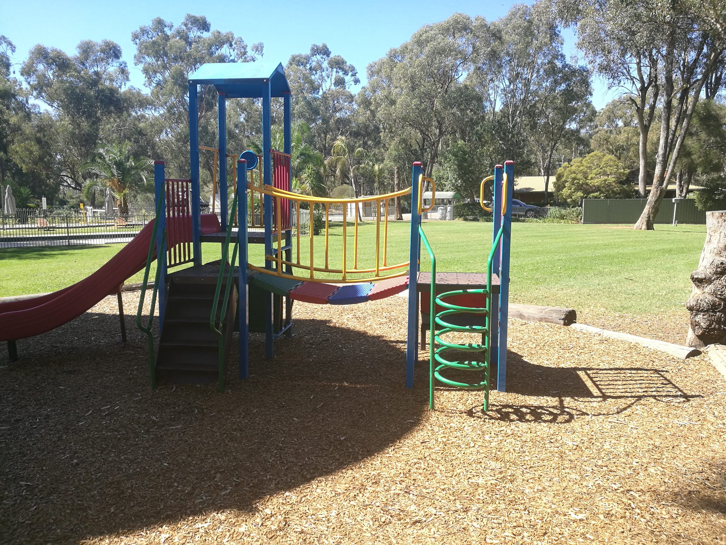 Colourful playground tower with red slide and spiral steps at Torrumbarry Weir Holiday Park, set on bark chips near the pool and lawn
