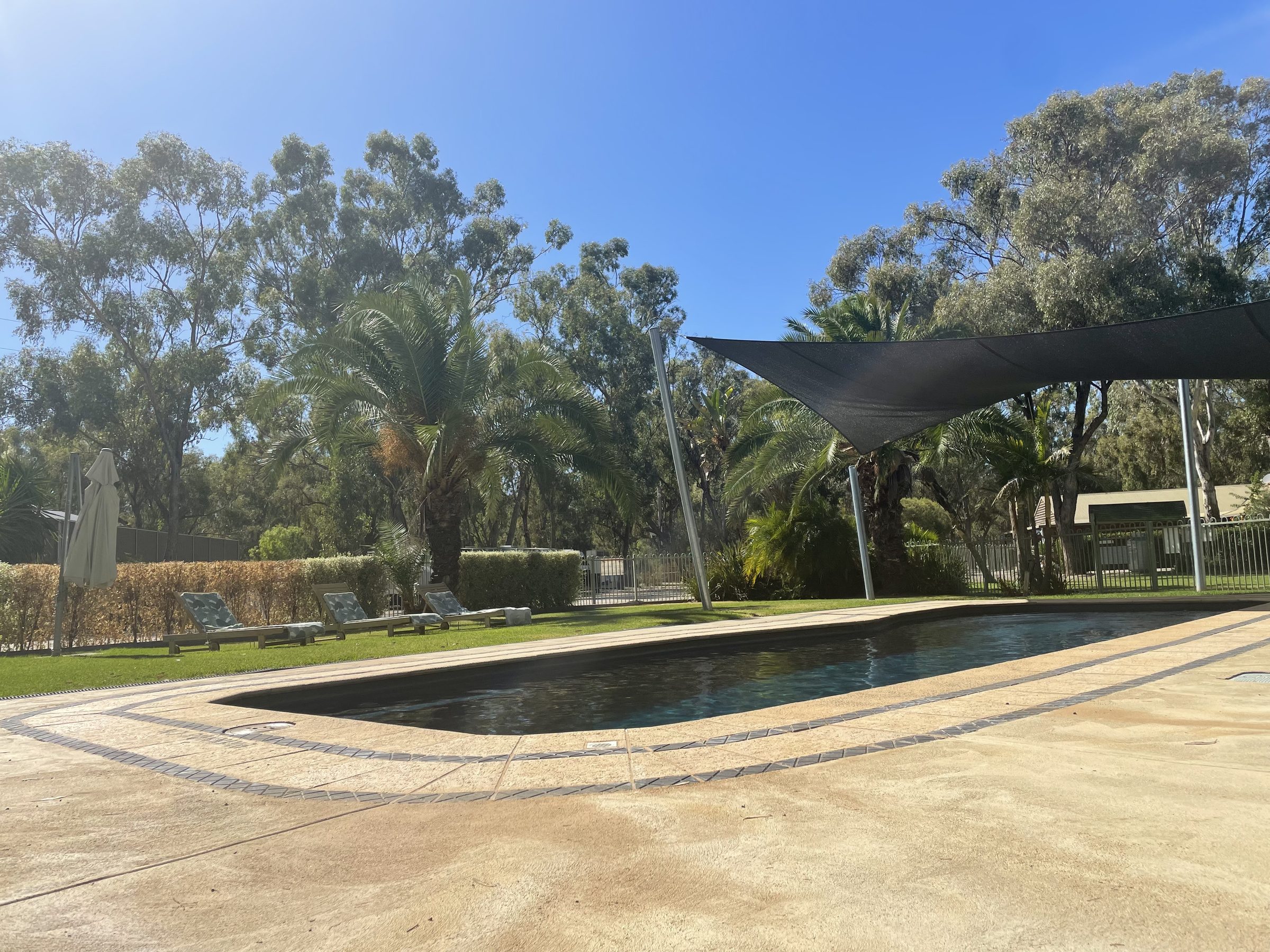 Swimming pool with shade sail and palms under blue sky at Torrumbarry Weir Holiday Park