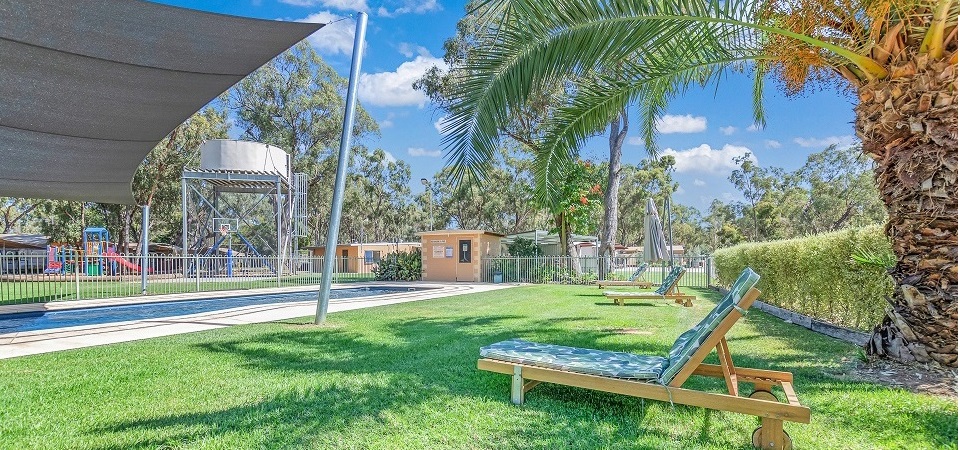 Sun loungers on green lawn beside the fenced swimming pool at Torrumbarry Weir Holiday Park, with shade sail and palm tree