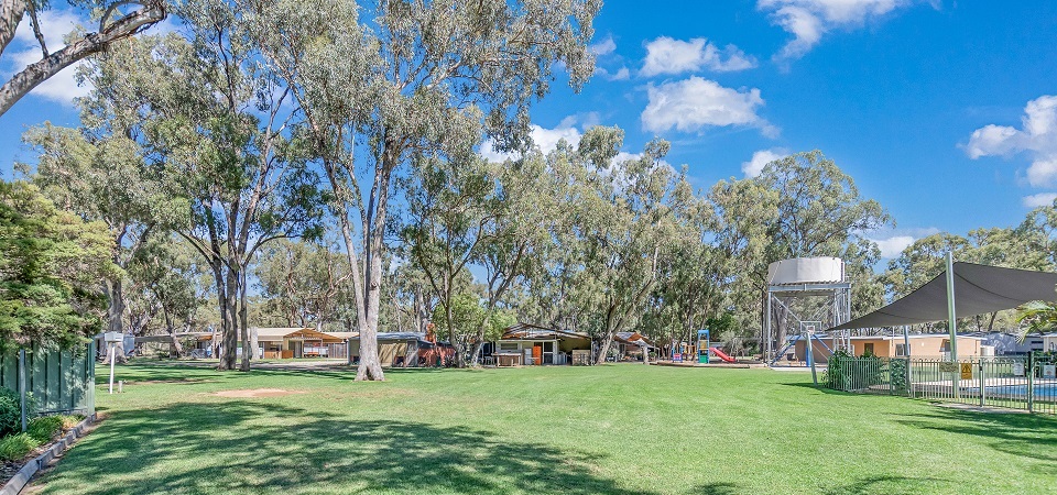 Wide green lawn beneath gum trees at Torrumbarry Weir Holiday Park, with playground, pool shade sail and cabins in Patho VIC
