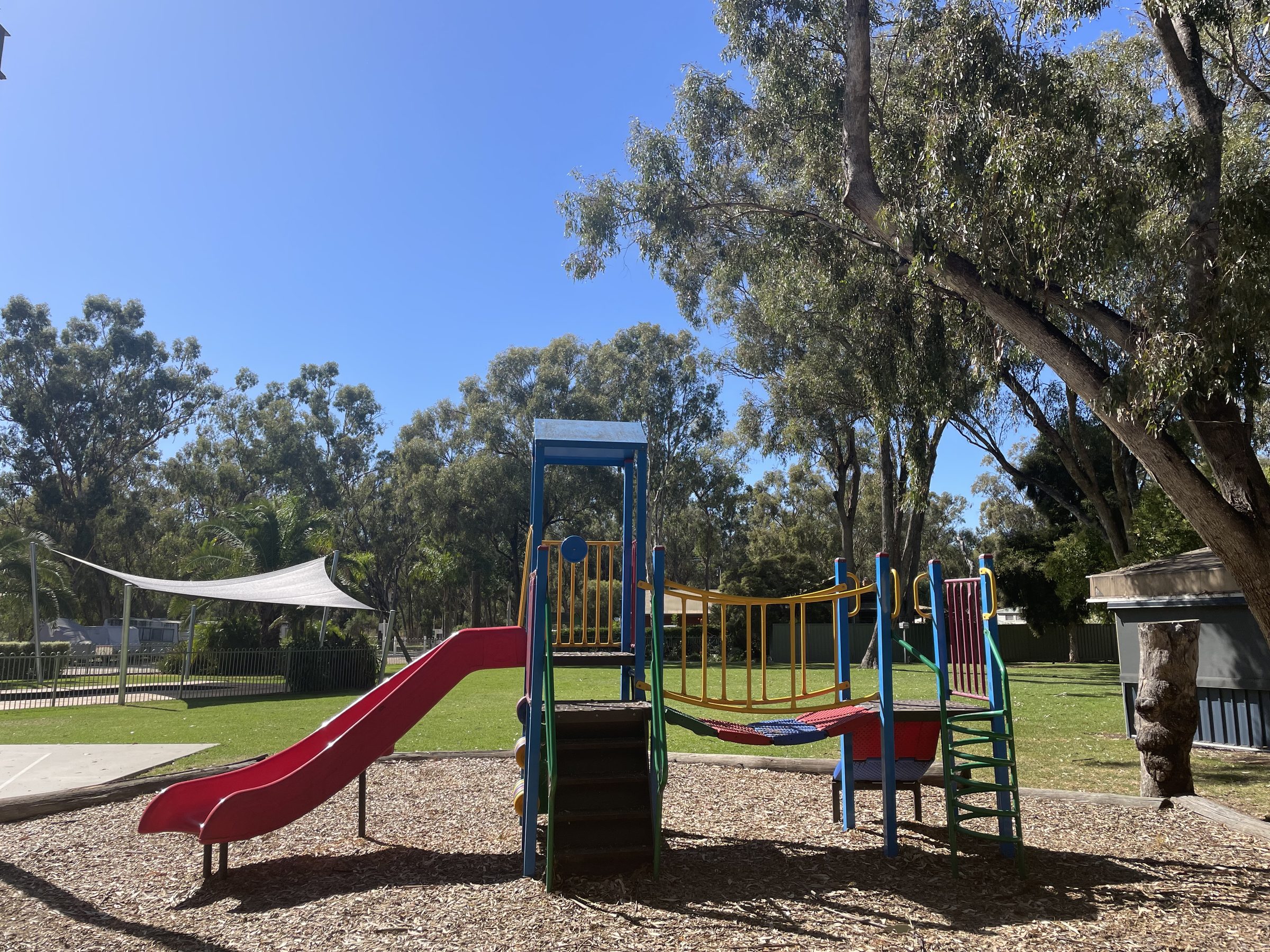 Red twin slide and climbing frame on woodchip playground under shady gum trees at Torrumbarry Weir Holiday Park, Patho VIC