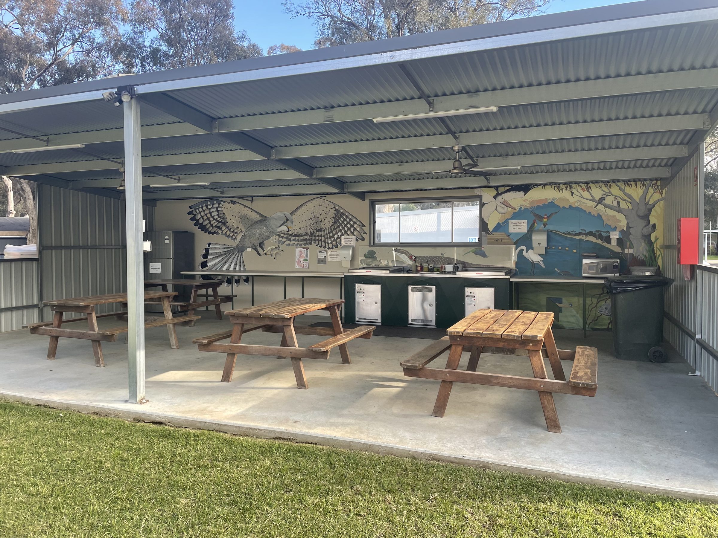 Open-sided camp kitchen shelter at Torrumbarry Weir Holiday Park with wooden picnic tables, hotplates and native bird murals