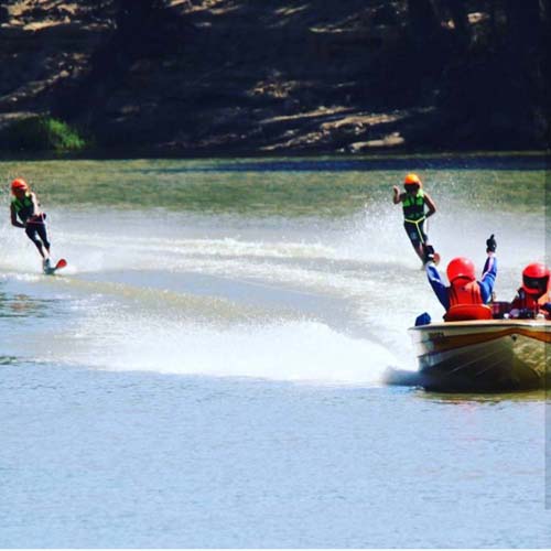 Two water skiers in helmets and life jackets skiing behind a boat on the river at Torrumbarry Weir Holiday Park