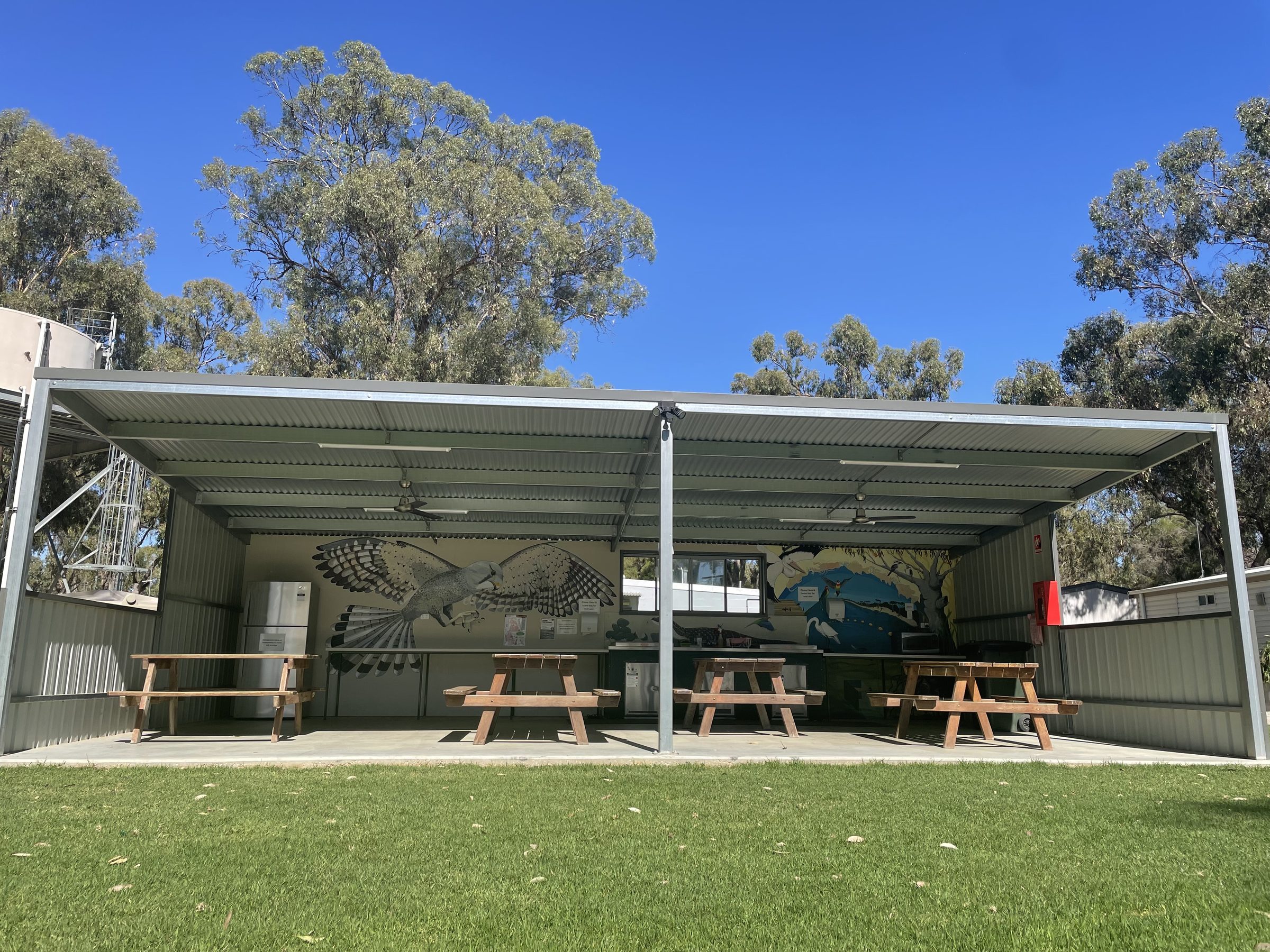 Open-sided camp kitchen with bird murals, four picnic tables and stainless fridges at Torrumbarry Weir Holiday Park caravan park, Patho VIC