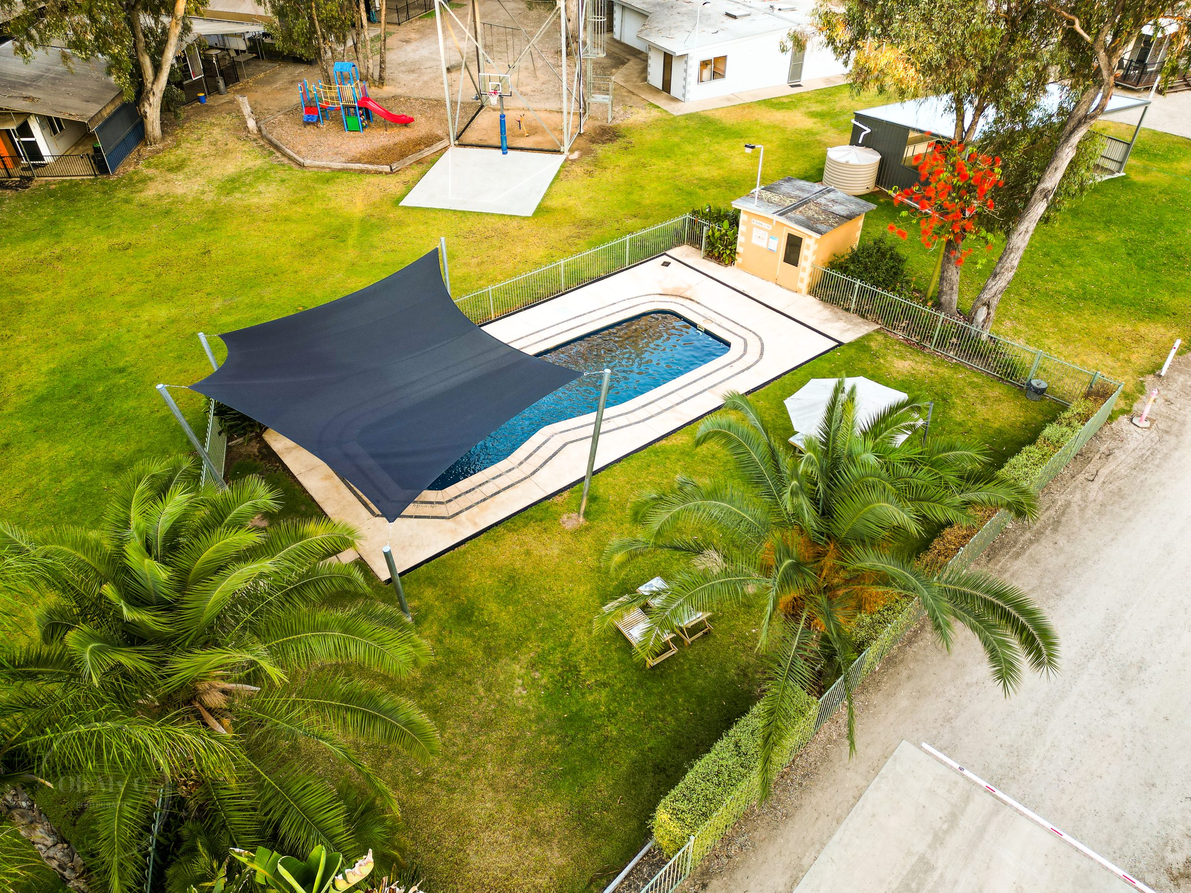 Aerial view of the fenced swimming pool with navy shade sail, palm trees and lawn at Torrumbarry Weir Holiday Park caravan park
